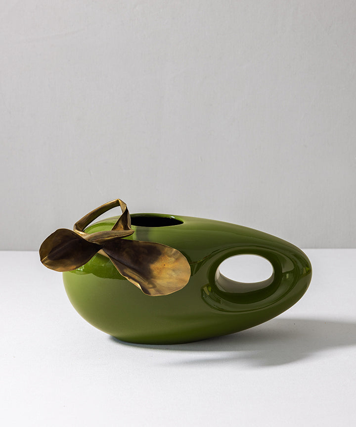 An abstract olive green ceramic vase with a metallic gold leaf-shaped spout and a side cutout, set against a plain light gray background.