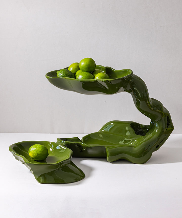 A glossy dark green sculptural ceramic fruit stand shaped like twisted branches or leaves, holding several green citrus fruits, photographed against a plain light gray studio background.