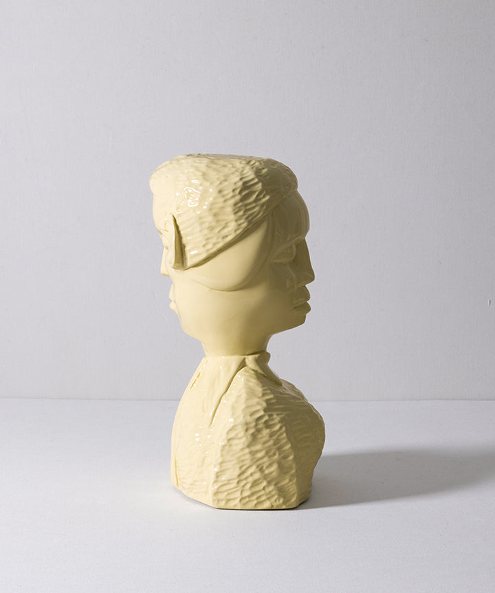 A pale yellow sculptural bust sculpture of two overlapping human heads and shoulders with a textured, ribbed surface, photographed against a neutral light gray studio background.