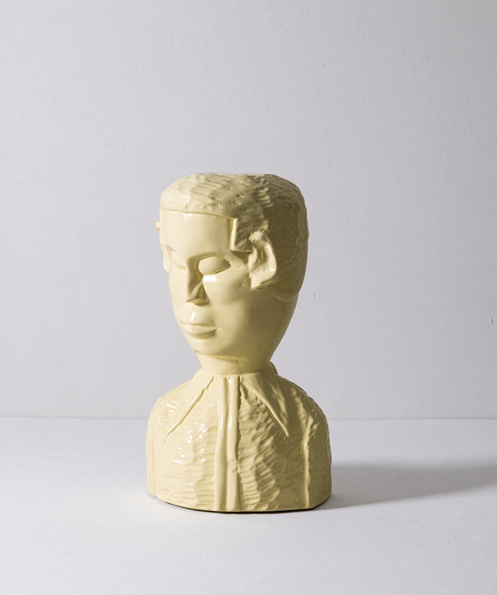A pale yellow sculptural bust of a face with short hair and a textured collared garment, photographed against a plain light gray studio background.