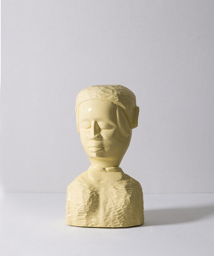 A small sculptural bust of a face with closed eyes, made of glossy pale yellow ceramic, set against a plain light gray background.