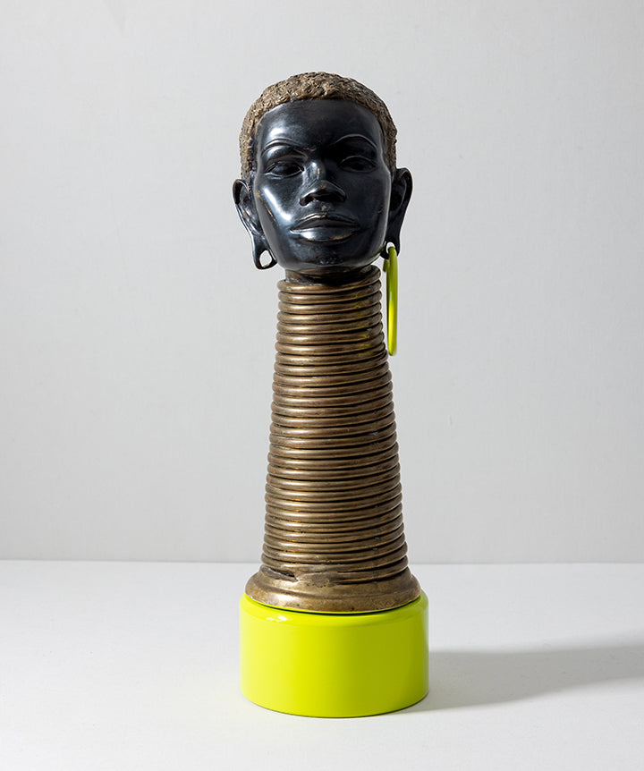 A sculptural decorative object featuring a glossy black human head with short textured hair, mounted on a ribbed metallic gold neck, with a bright yellow-green cylindrical base and matching earring, photographed against a plain light gray studio background.