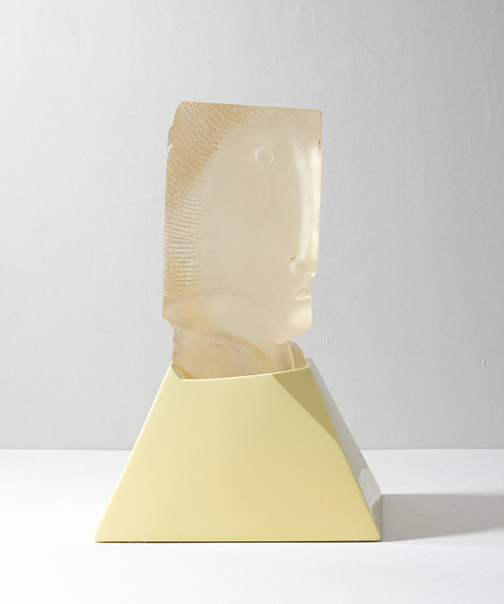 A decorative sculpture featuring a semi-translucent cast glass beige carved face emerging from a matte pale yellow geometric pedestal, set against a neutral light gray background.