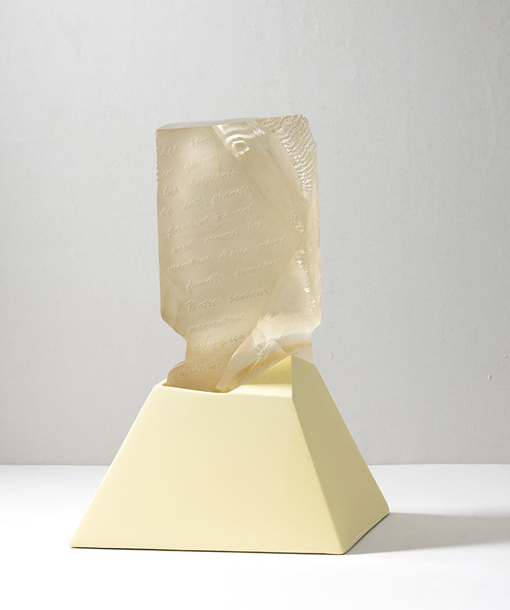 A decorative sculpture featuring a semi-translucent cast glass beige carved face emerging from a matte pale yellow geometric pedestal, set against a neutral light gray background.