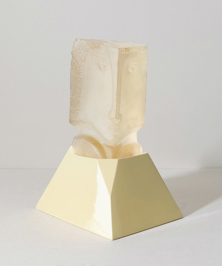 A decorative sculpture featuring a semi-translucent cast glass beige carved face emerging from a matte pale yellow geometric pedestal, set against a neutral light gray background.