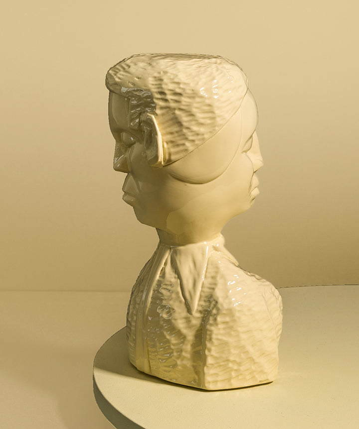 A glossy sculptural bust hand carved wood double sided brutalist bust of man and woman., shown on a round pedestal against a warm beige studio background.