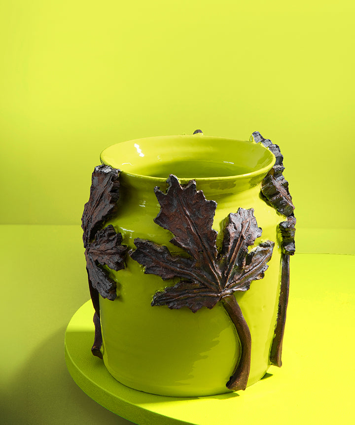 A bright lime green ceramic planter pot decorated with three dark brown leaf and stem sculptures, set against a matching lime green background.