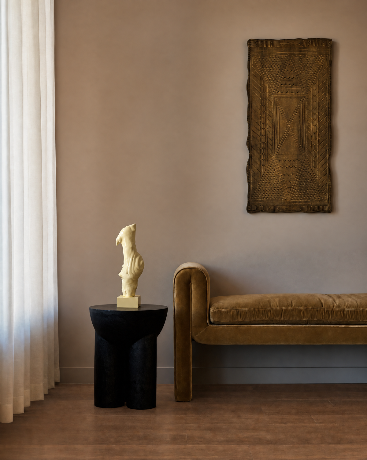 A styled interior scene with a brown velvet bench against a warm beige wall, a black side table with a cream sculptural hand carved wood object reads like a ballet slipper, and a large textured brown wall hanging above the bench.