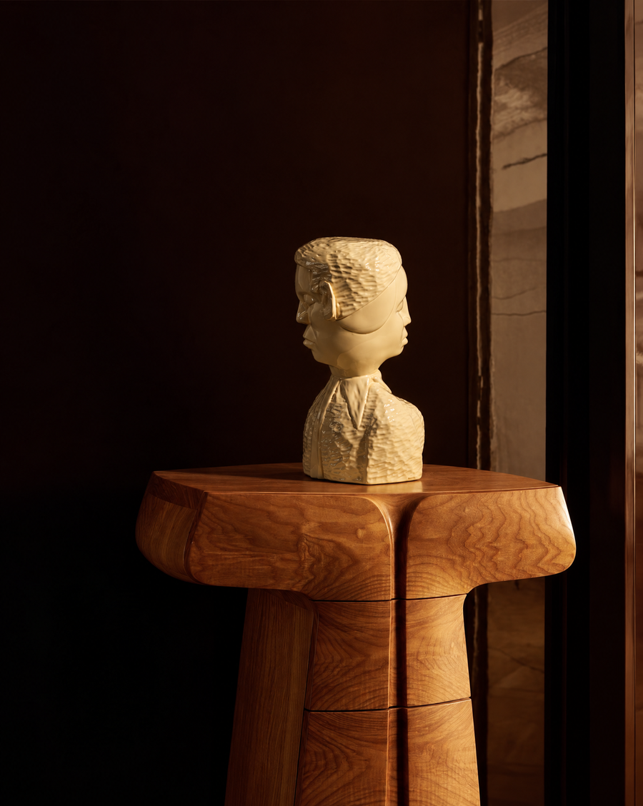 Hand carved wood double sided brutalist bust of man and woman on a wooden pedestal with a dark burgundy wall behind it.
