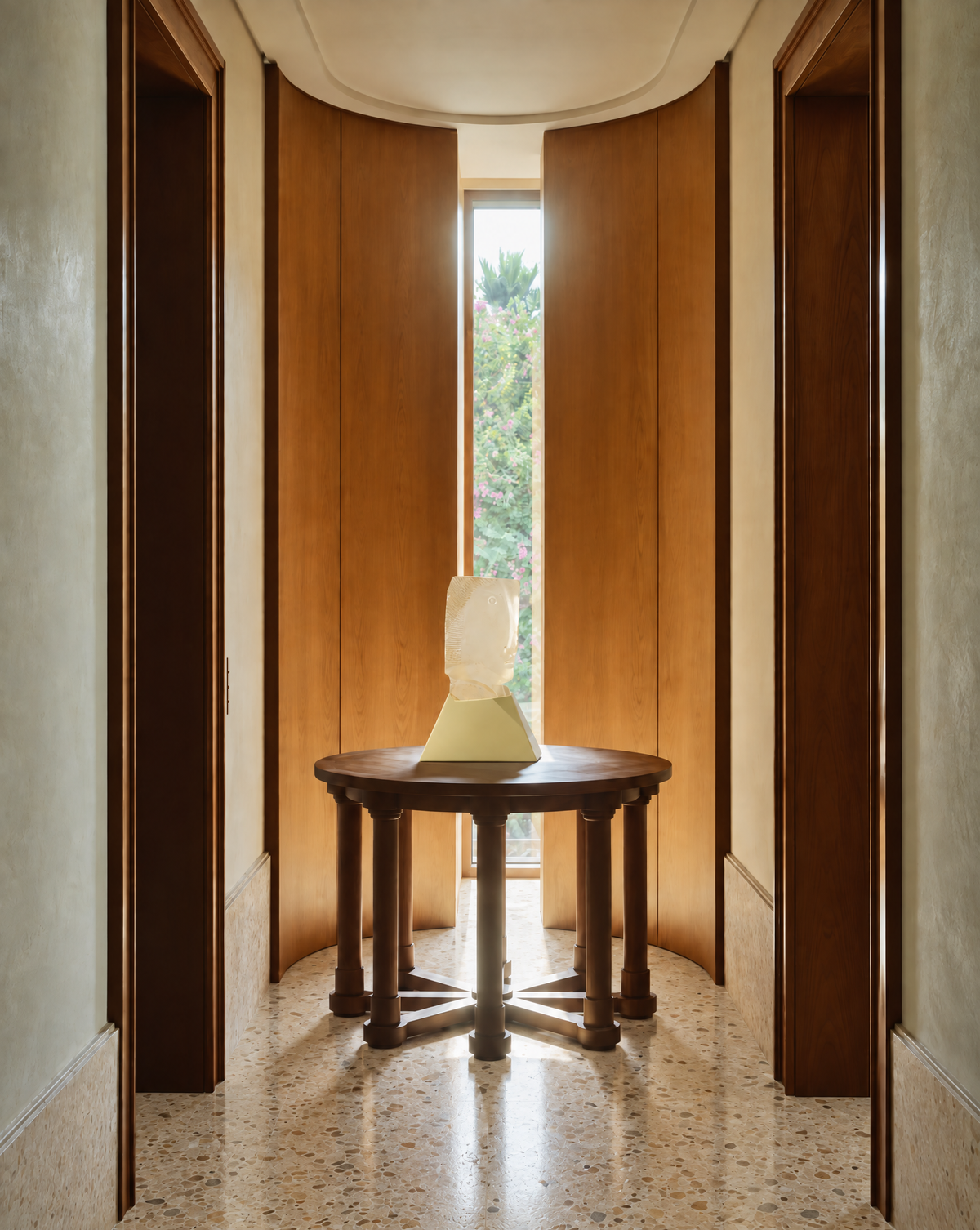A round dark wood pedestal table with four legs stands in a narrow hallway with curved wooden doors and a window at the end, holding this signed cast glass Tête sculpture by French artist Gilles Chabrier.