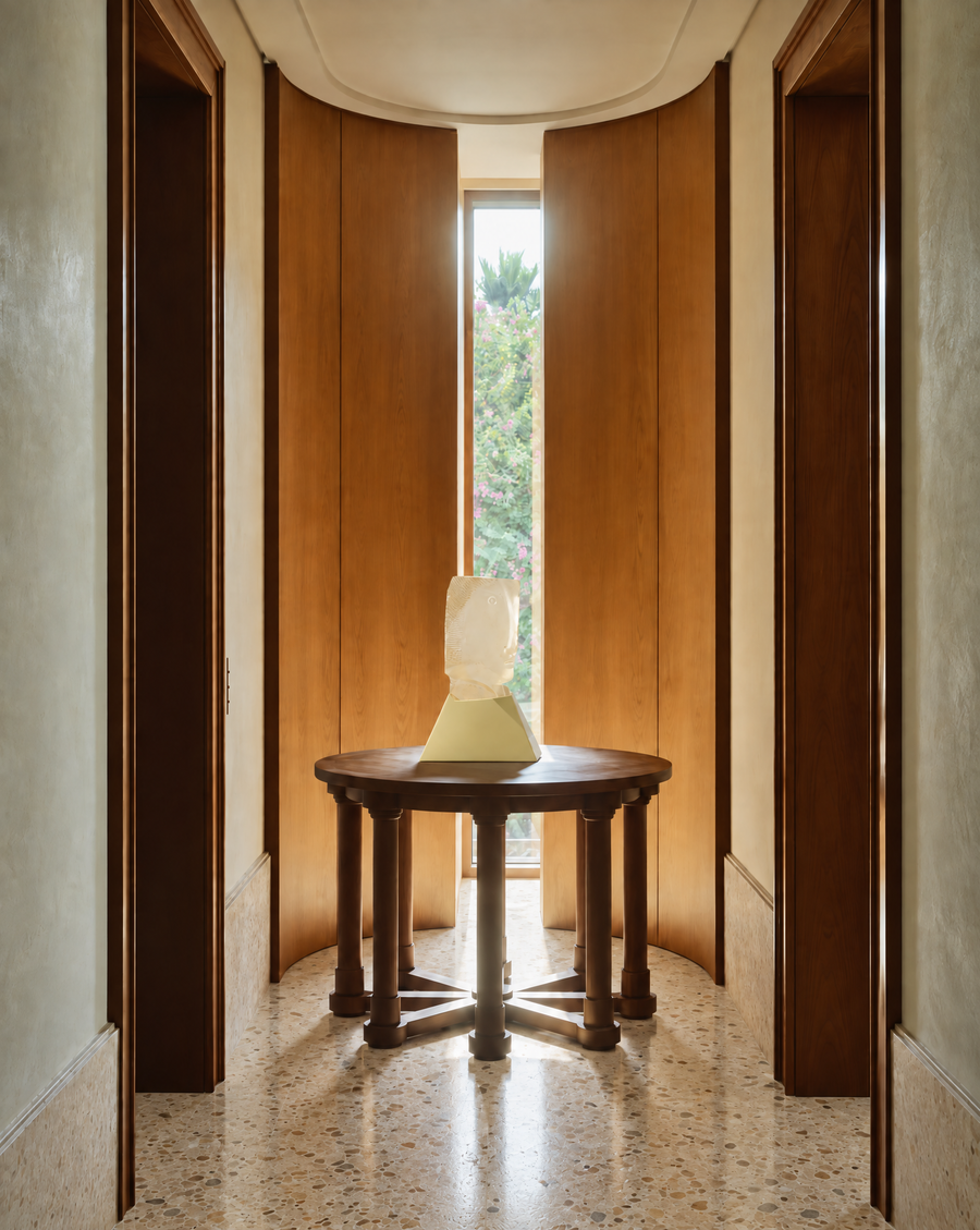A round dark wood pedestal table with four legs stands in a narrow hallway with curved wooden doors and a window at the end, holding this signed cast glass Tête sculpture by French artist Gilles Chabrier.