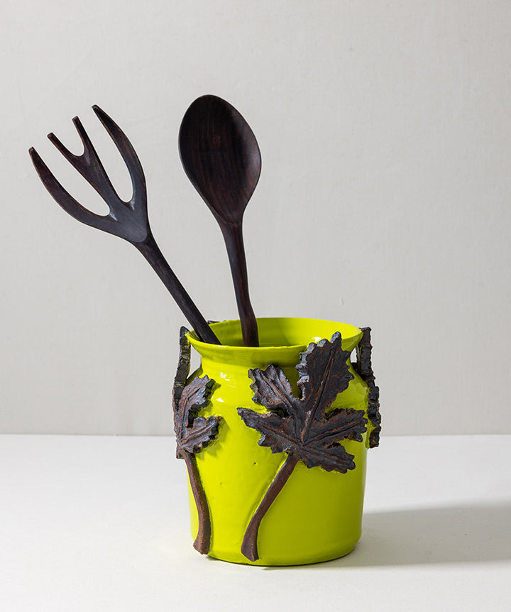 A bright lime green ceramic utensil holder with dark brown leaf and handle details, holding two dark wooden kitchen utensils, a spoon and a fork, against a plain light gray background.