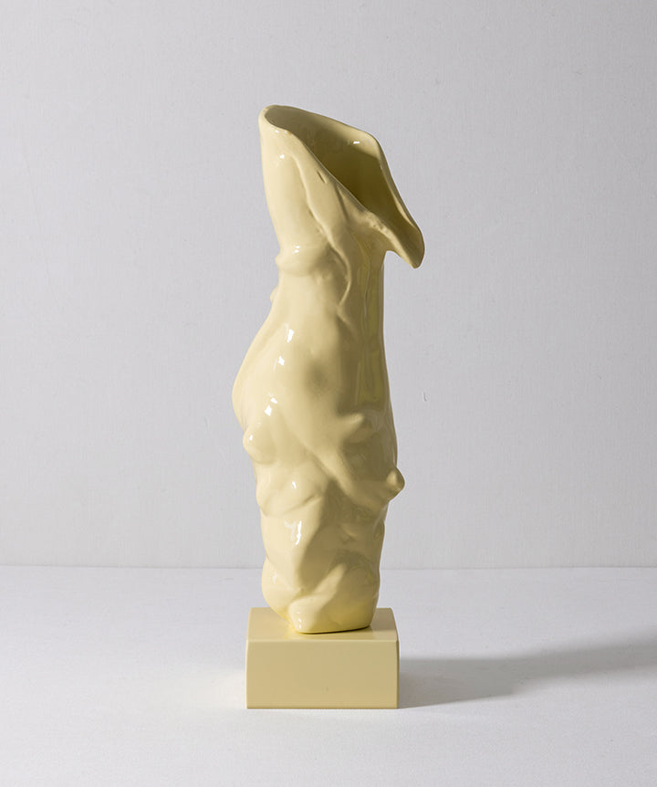 A glossy pale yellow hand carved wood object reads like a ballet slipper vase with an abstract, draped organic form, displayed on a matching rectangular pedestal against a plain light gray studio background.