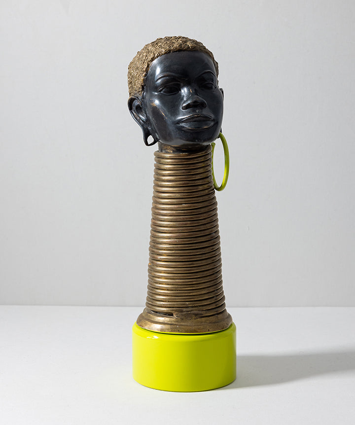 A sculptural decorative object featuring a glossy black human head with short textured hair, a ribbed metallic neck in a bronze/gold tone, and a bright yellow base and hoop earring, photographed against a plain light gray studio background.