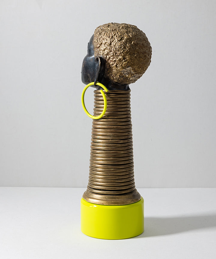 A sculptural decorative object with a textured metallic gold head, a ribbed cylindrical body, a bright yellow base, and a large black earring with a yellow hoop, photographed against a plain light gray studio background.