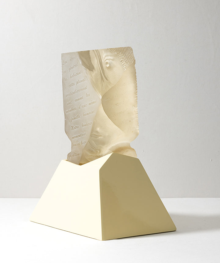 A decorative sculpture featuring a semi-translucent cast glass beige carved face emerging from a matte pale yellow geometric pedestal, set against a neutral light gray background. 