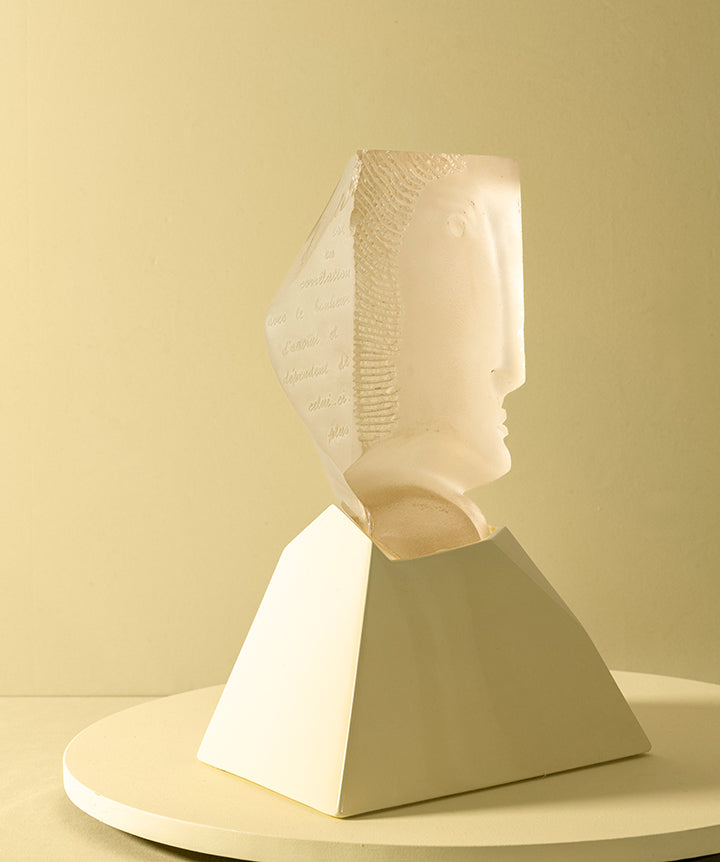 A decorative sculpture featuring a semi-translucent cast glass beige carved face emerging from a matte pale yellow geometric pedestal, set against a cream yellow background. 