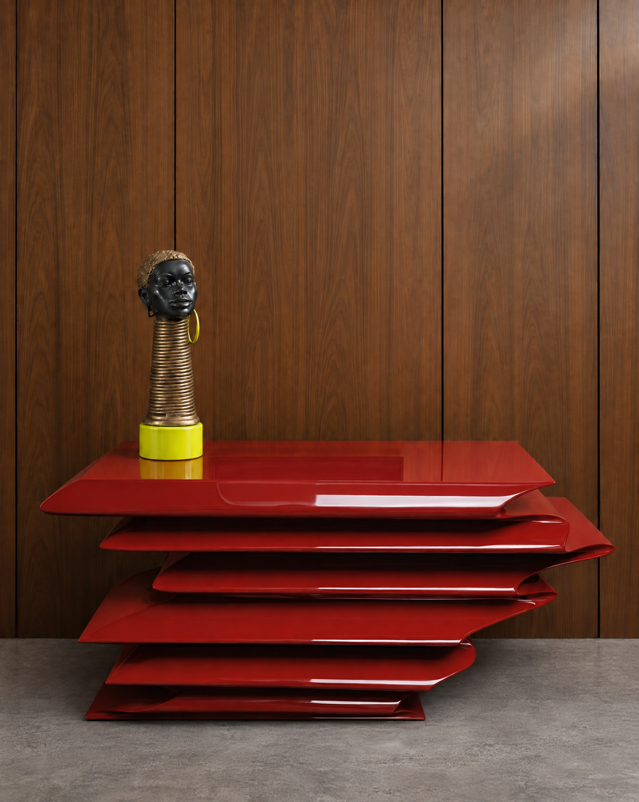 A glossy red sculptural side table with layered, wavy rectangular plinth-style tiers, shown against a dark wood-paneled wall on a gray floor. A regal celebration of African beauty. Her oversized hoop was dismantled and base was filled, both to be coated in a neon pop that would allow her brass body to shine. Figure sits atop a solid marble base also coated in Wilson.