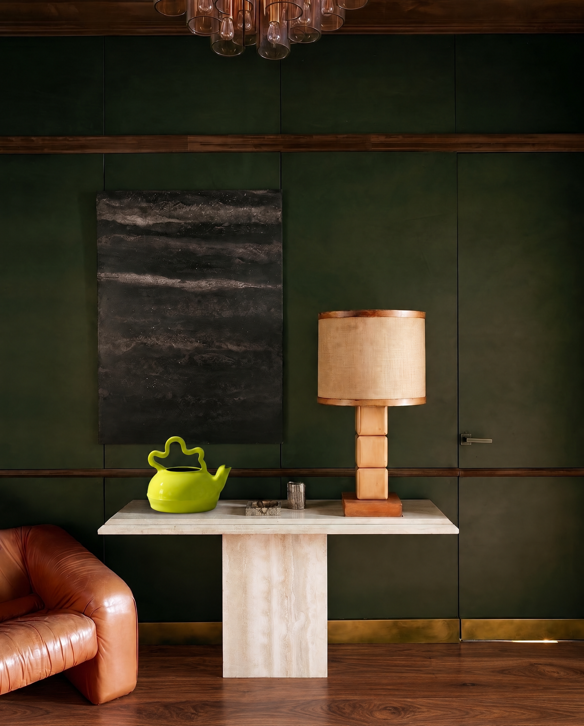 A styled interior scene with a dark green paneled wall, a light wood console table, a floor lamp with a beige drum shade, a bright green teapot, and part of a brown leather armchair visible in the lower left.